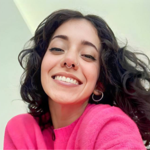 Venessarey smiles brightly in a pink sweater, her dark curly hair framing her face, with a nose ring adding a playful touch to her cheerful and vibrant look.